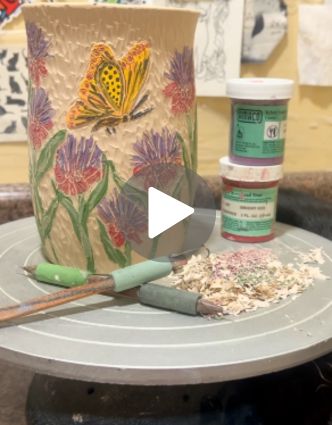 A textured ceramic vase featuring hand-carved and painted zinnias and a vibrant yellow butterfly sits on a pottery wheel, surrounded by carving tools, colorful underglaze jars, and clay shavings.