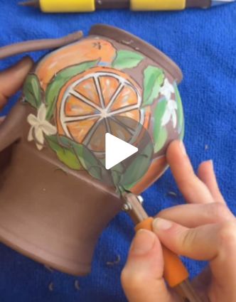 A hand uses a FP0 tool to carve fine details into an orange slice design on a painted ceramic mug, surrounded by additional pottery tools on a blue cloth backdrop.