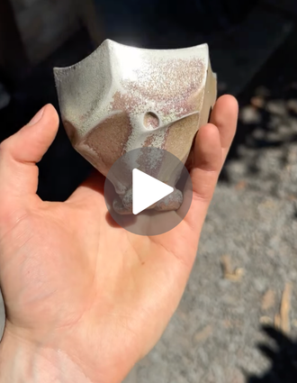 Close-up of a faceted ceramic bowl fragment with a shimmering wood-fired glaze, held in the artist's hand outdoors.