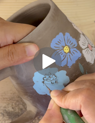 Artist using a DiamondCore® L1 sgraffito stylus to carve fine floral details into blue underglaze on a leather-hard mug.