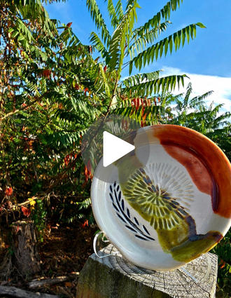 A spoon rest and garlic grater duo with leafy carvings and earthy glazes rests against a weathered tree stump, framed by vibrant fall foliage. The colors echo the reds, greens, and golds of autumn.