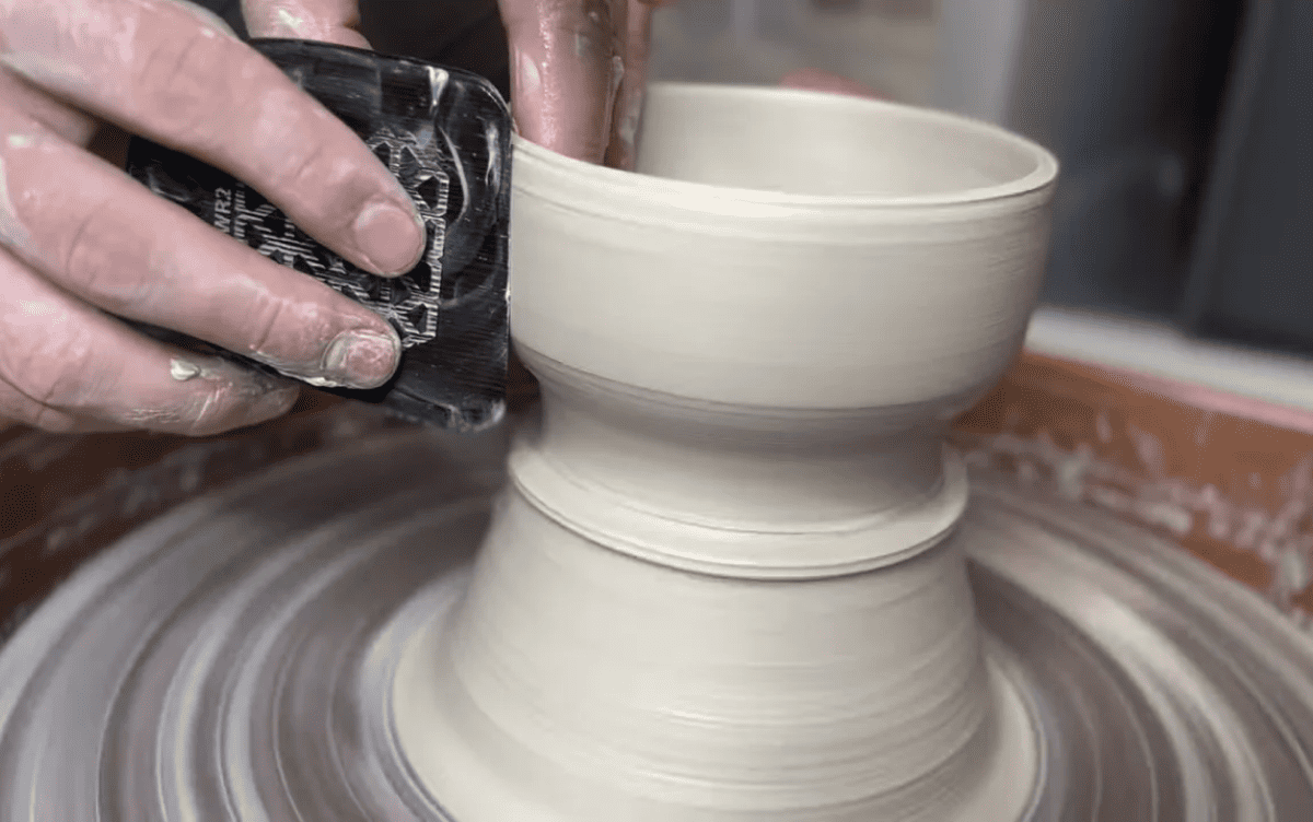 A potter uses a DiamondCore Tools trimming tool to refine the shape of a clay vessel on a spinning pottery wheel, smoothing the surface with precise control.