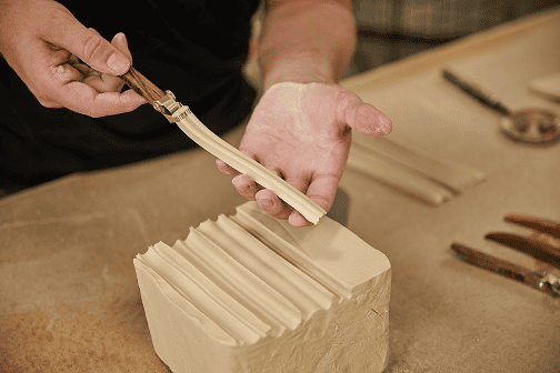 A ceramic artist uses a DiamondCore Tools carving tool to slice a smooth, ribbon-like strip from a block of clay, revealing multiple crisp, parallel carved lines.