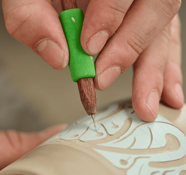 A close-up of a person carving a detailed pattern into a clay surface using a fine-tipped DiamondCore carving tool with a green grip.
